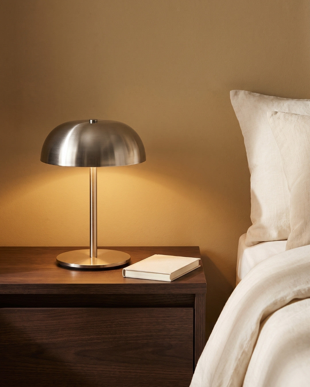 Gavetti table lamp in silver lit on a nightstand beside a book
